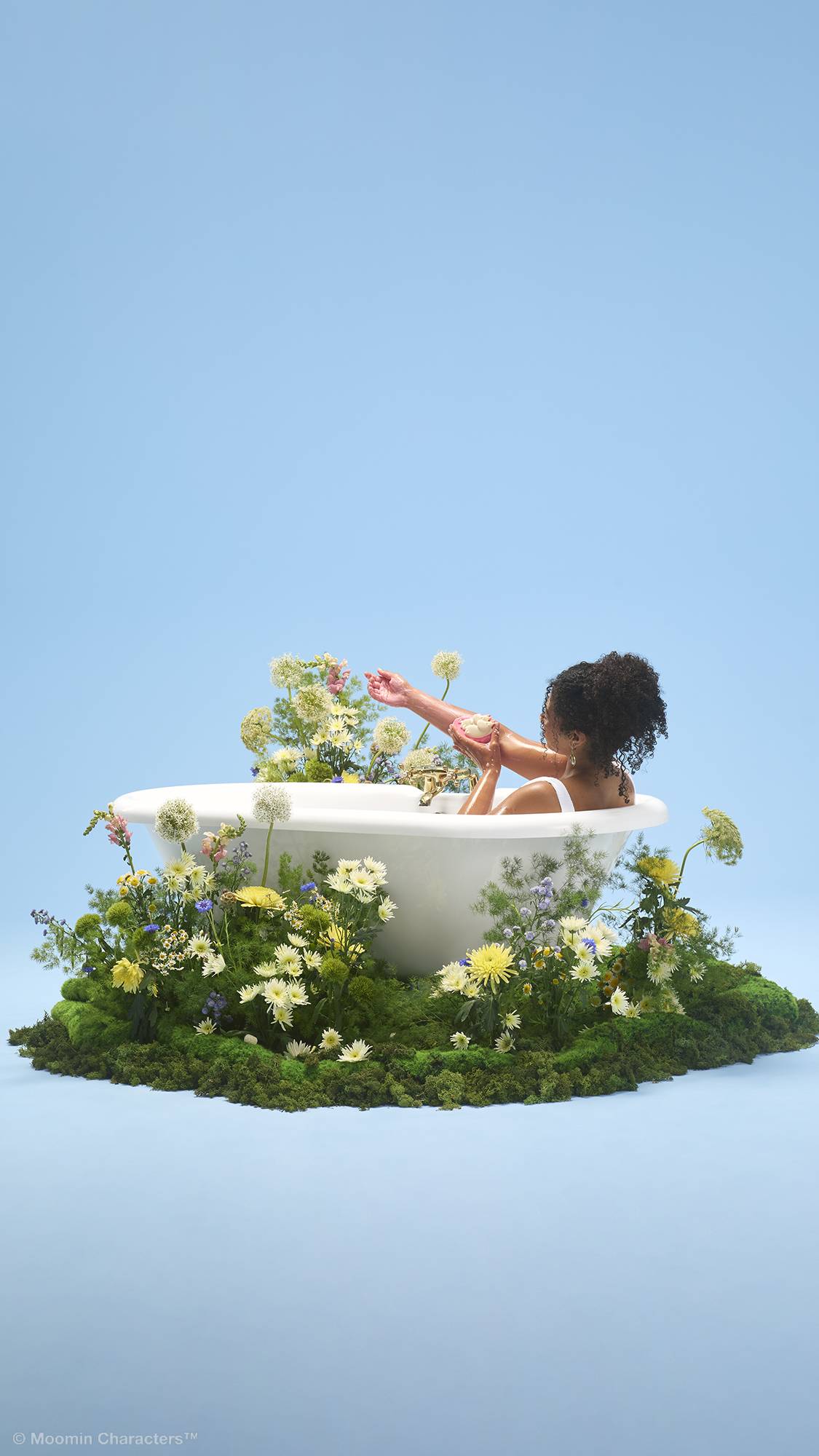A person with curly hair sits in a white bathtub, washing their arm with a pink and white "Moomin Delight" soap and body wash. The tub sits on a patch of green moss filled with wildflowers, set against a plain light blue background.