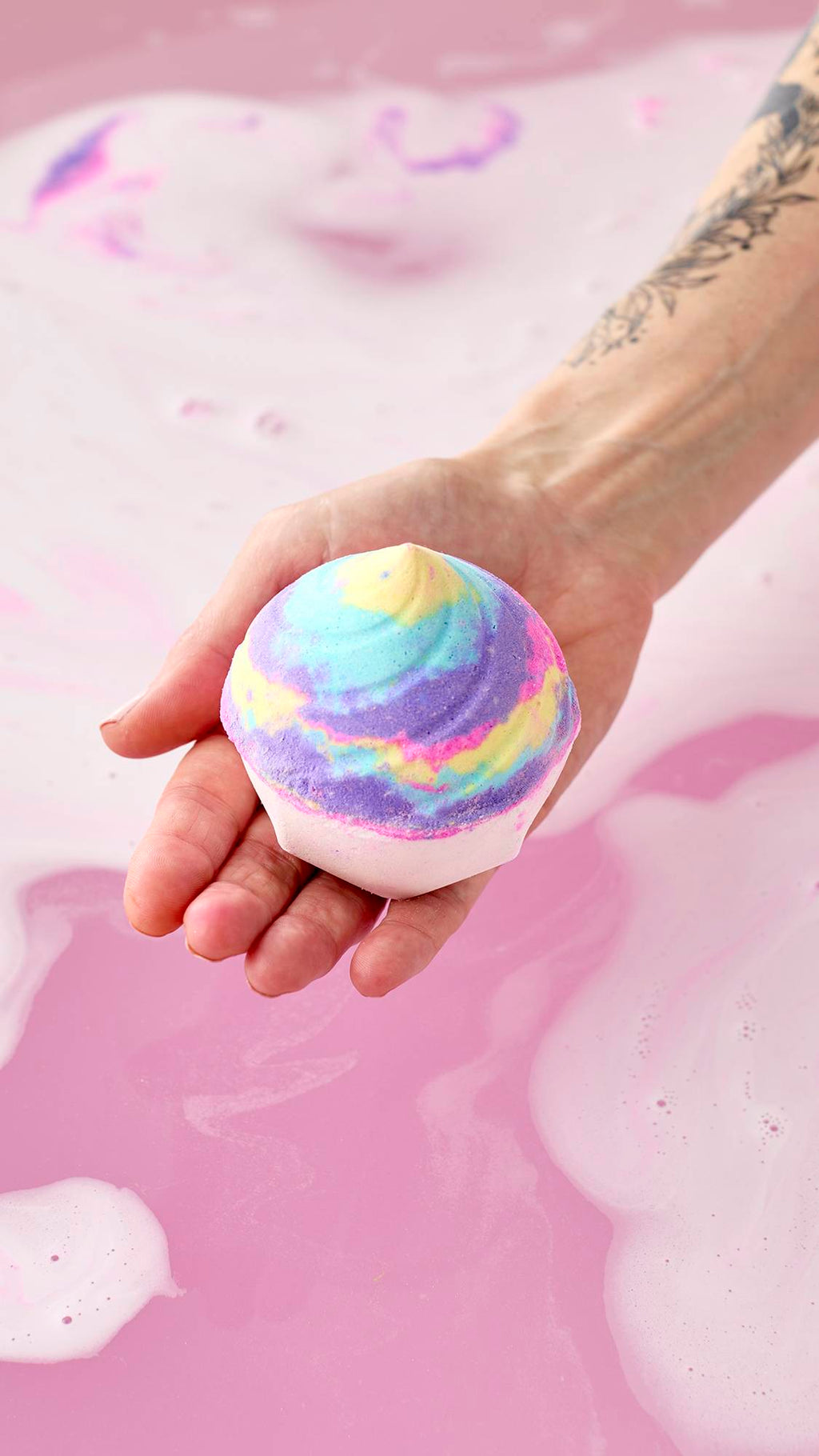 A hand holds the Unicorn Poop Bath Bomb over a bathtub filled with gently swirling pink and purple water. The whimsical, swirl-shaped bath bomb features a white base topped with a vibrant mix of pastel blue, yellow, purple, and pink hues.