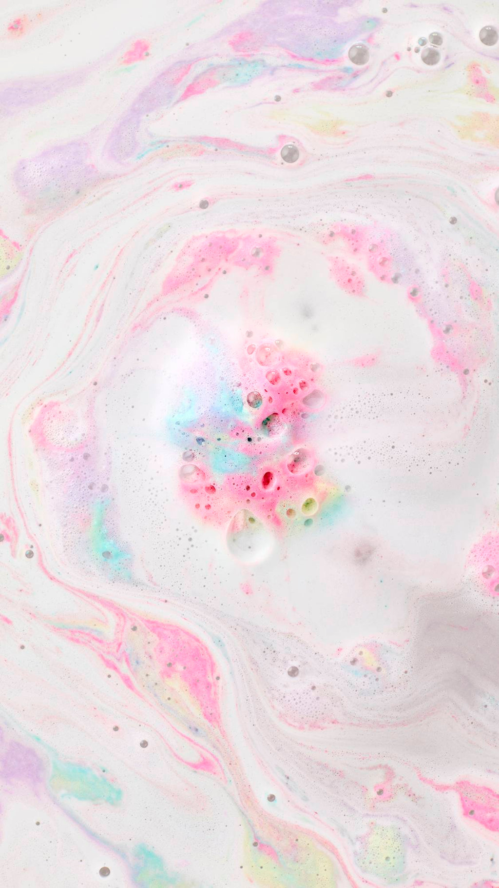 A top-down view of milky white bathwater with pastel swirls of pink, yellow, and turquoise foam. In the center, a cluster of vibrant pink and blue bubbles fizzes, creating a marbled "bath art" effect across the surface.