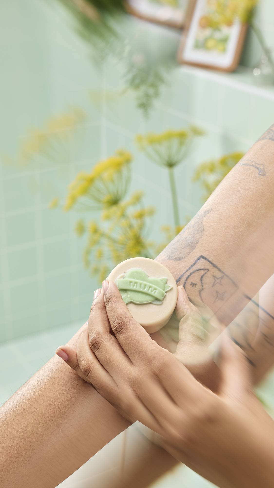 Hands massage a cream "Symbol of Love" oil bar with a green "MUM" banner onto a tattooed arm. Yellow flowers frame the scene against a mint green tiled background.