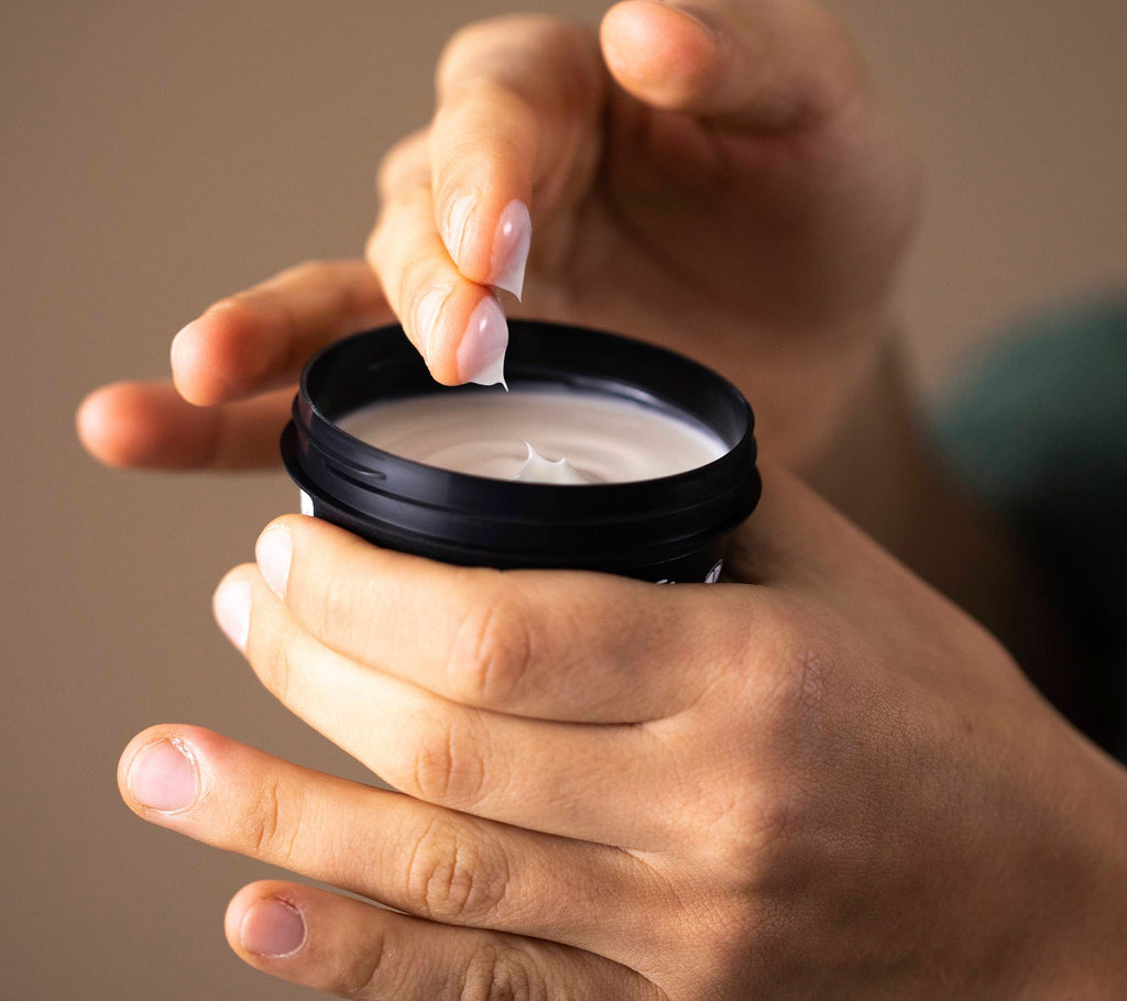 A close-up of hands holding the Cosmetic Lad pot with one hand dipping two fingers into the velvety, creamy moisturiser.