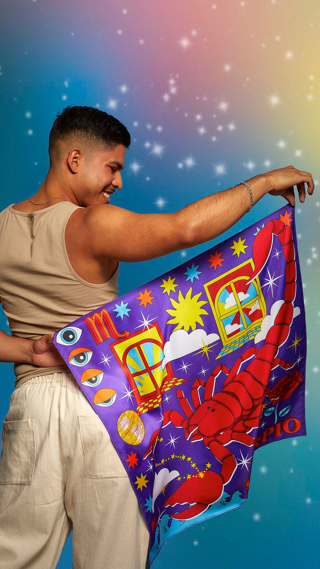 The image shows the model wearing a beige tank top and linen trousers. They are smiling on a pastel, sparkling background and holding the Scorpio knot wrap behind their back showing the full design.